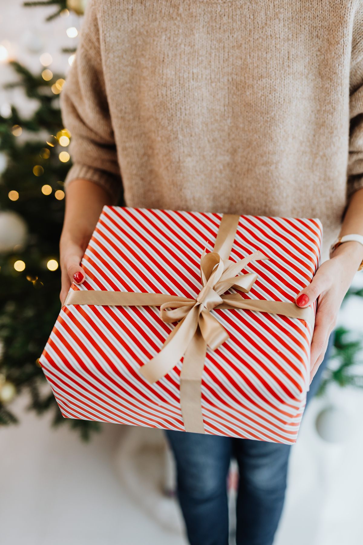 A close-up of a person holding a festive wrapped gift with red and white stripes near a decorated Christmas tree.